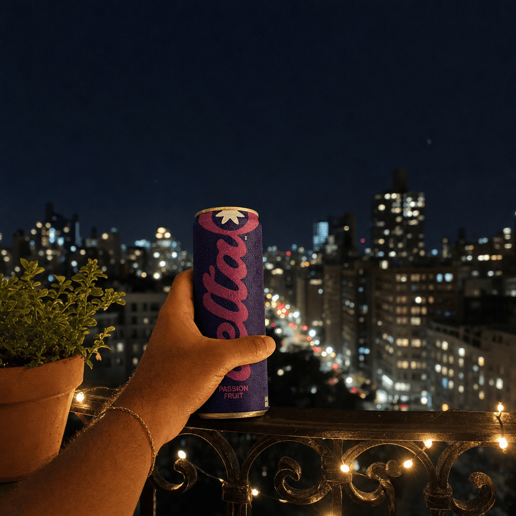A hand on a black wrought-iron balcony rail at night holding a 20mg Passion Fruit can, blurred city lights below