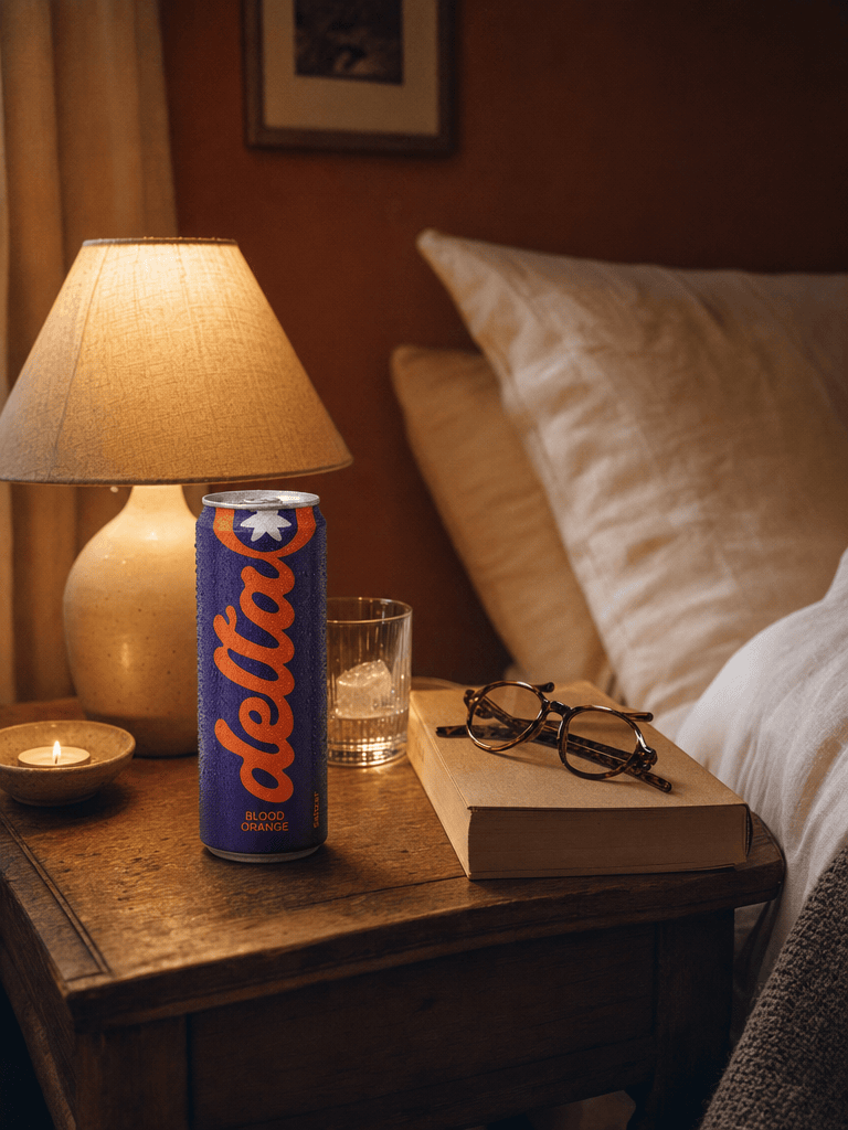 A bedside table at night with a 20mg Blood Orange can, paperback open face-down, glasses, dim warm bedside lamp